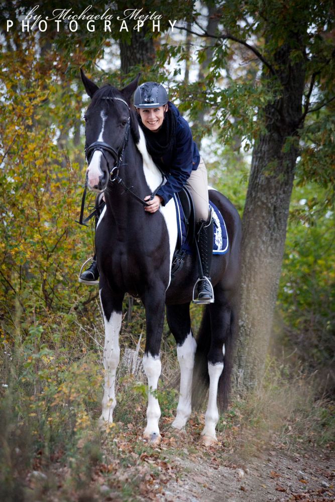 Smiling rider wearing a helmet on a black and white horse along a wooded trail in autumn colors to the left side of the path.