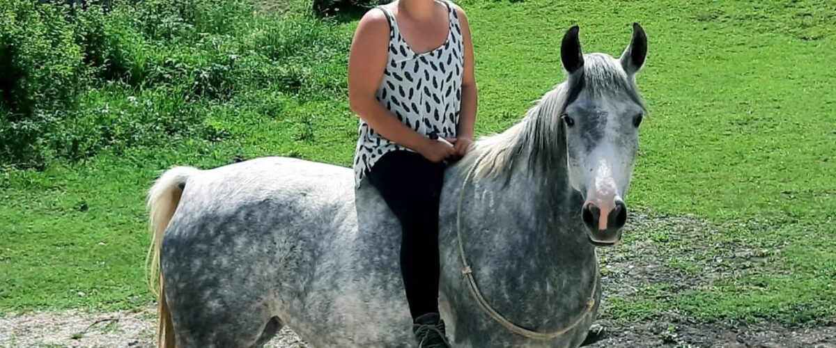 Woman wearing sunglasses sits astride a gray horse in a grassy paddock with wooden fences.