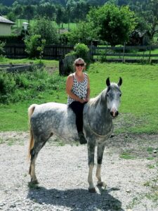 Woman wearing sunglasses sits astride a gray horse in a grassy paddock with wooden fences.