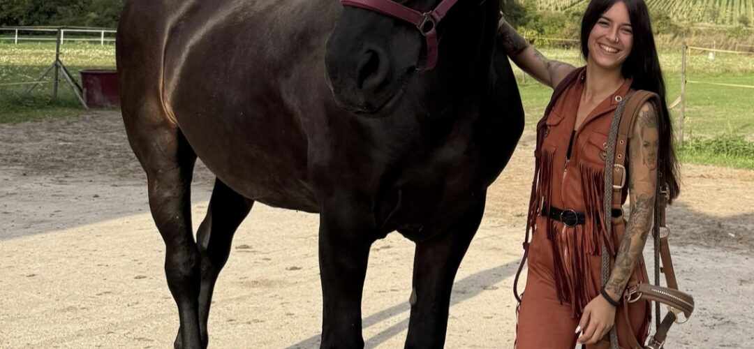 Smiling woman in a brown fringe outfit stands beside a large black horse in a sandy paddock.