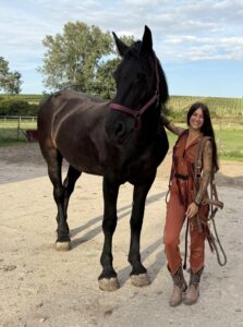 Smiling woman in a brown fringe outfit stands beside a large black horse in a sandy paddock.