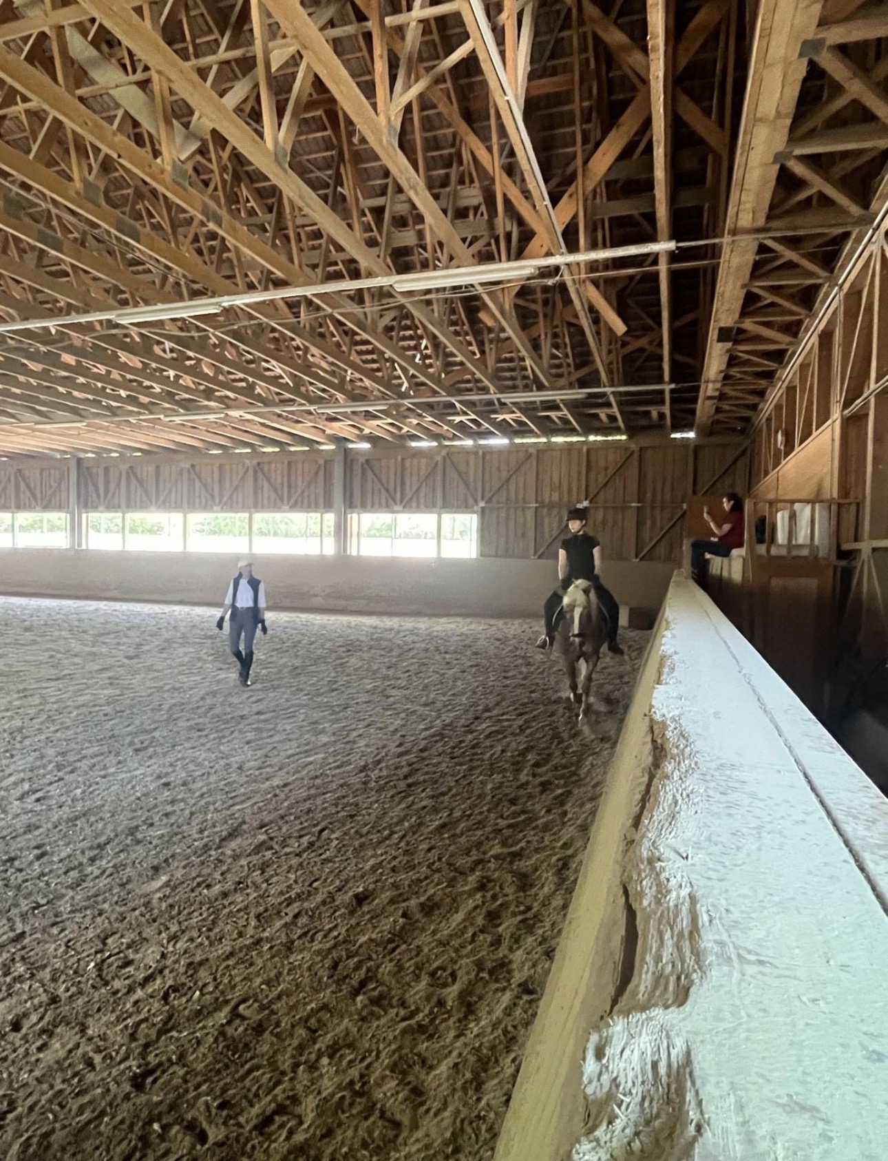 Indoor riding arena with exposed wooden trusses; a rider on a horse near the rail while a person walks across the sandy arena floor.